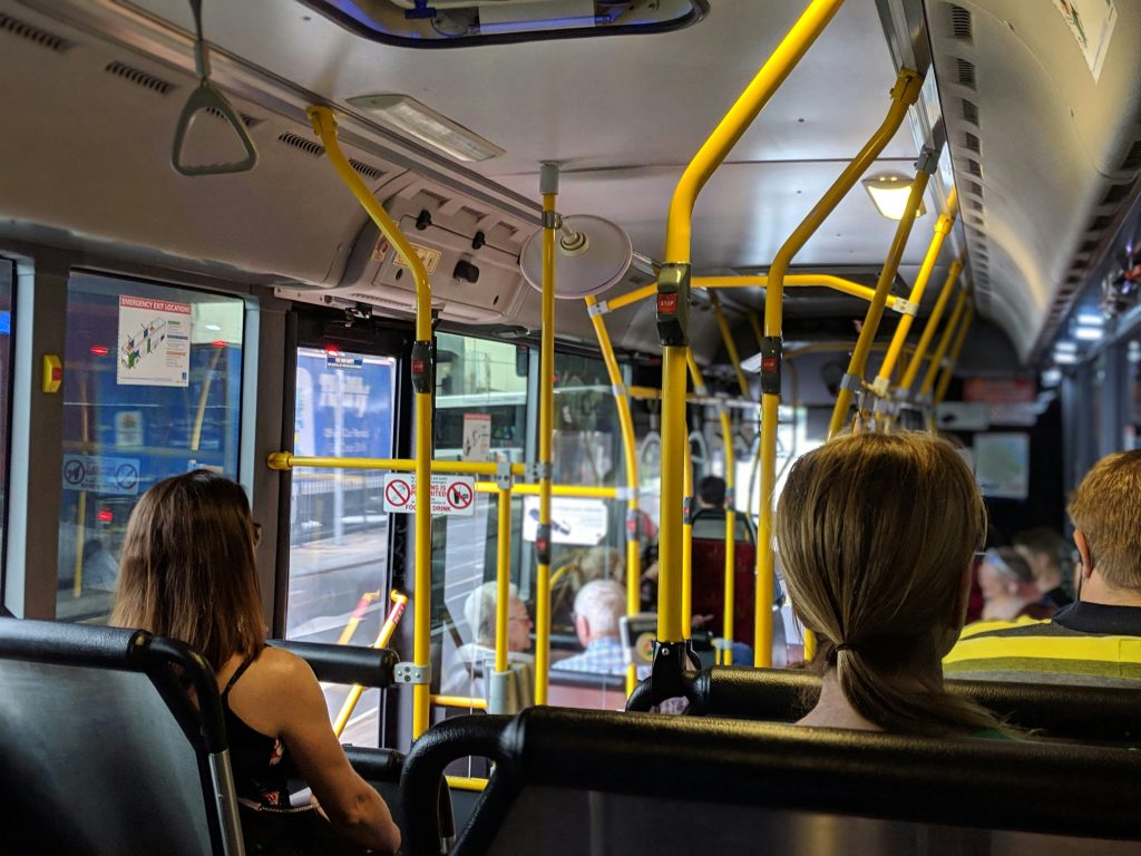 Image of commuters from behind seated on a bus with yellow railing via Unsplash